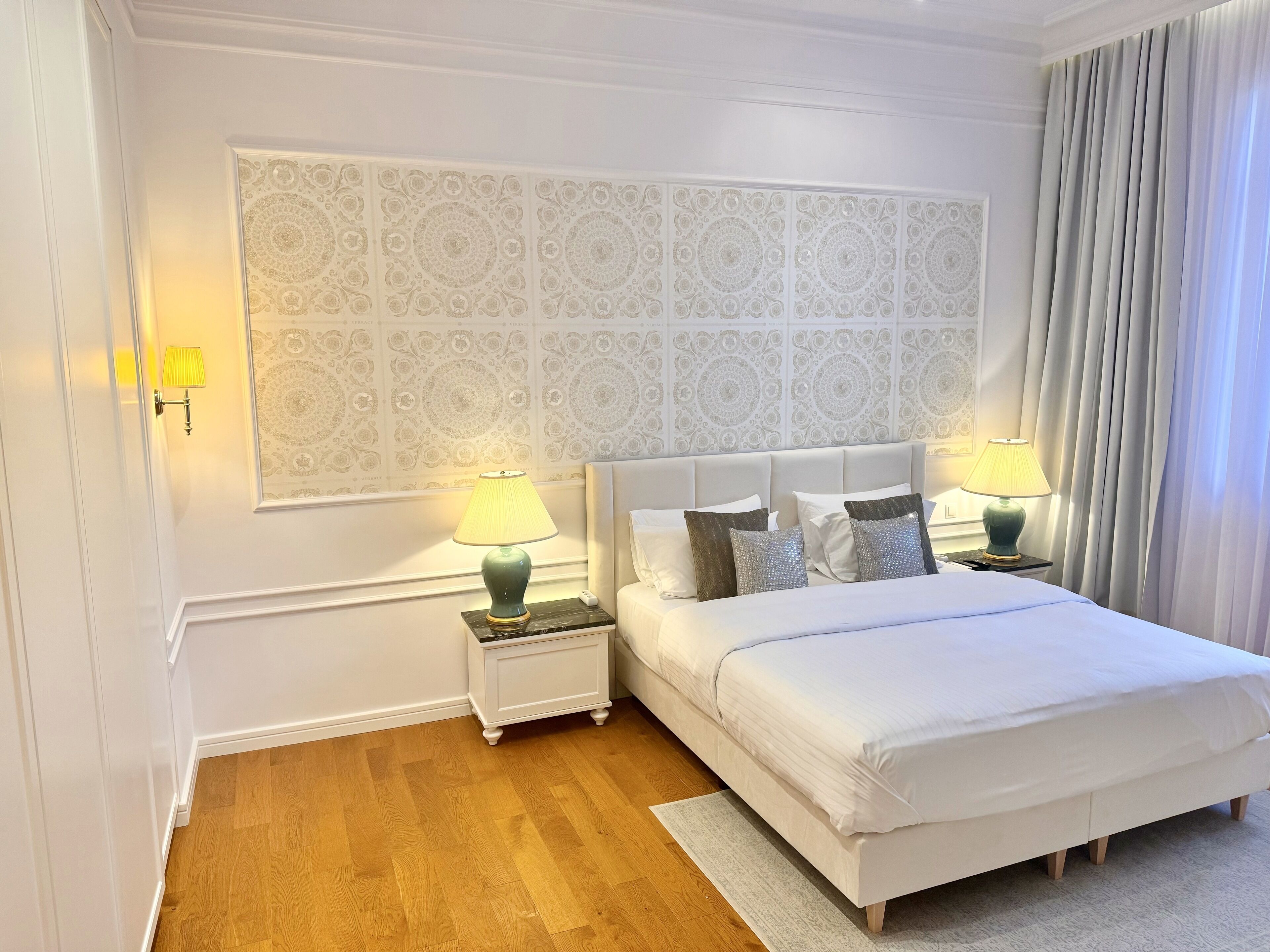 Een netjes ingerichte slaapkamer met een groot patroon op de muur, twee nachtkastjes met lampen, een bed met witte beddengoed en een houten vloer.