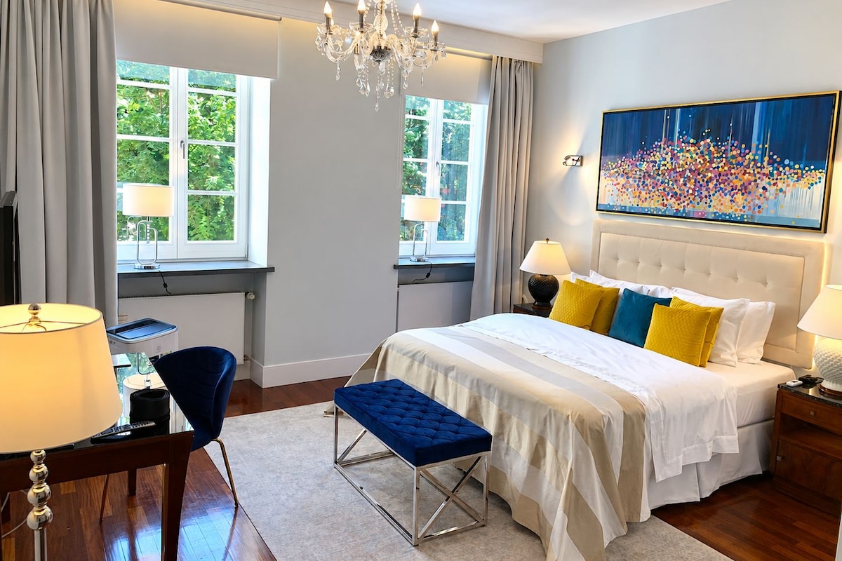 Ein Hotelzimmer mit einem großen Bett, zwei Nachttischlampen, einer Kandelaber, einem Fenster mit Vorhängen, einem Schreibtisch mit Stuhl und einem Bild an der Wand.