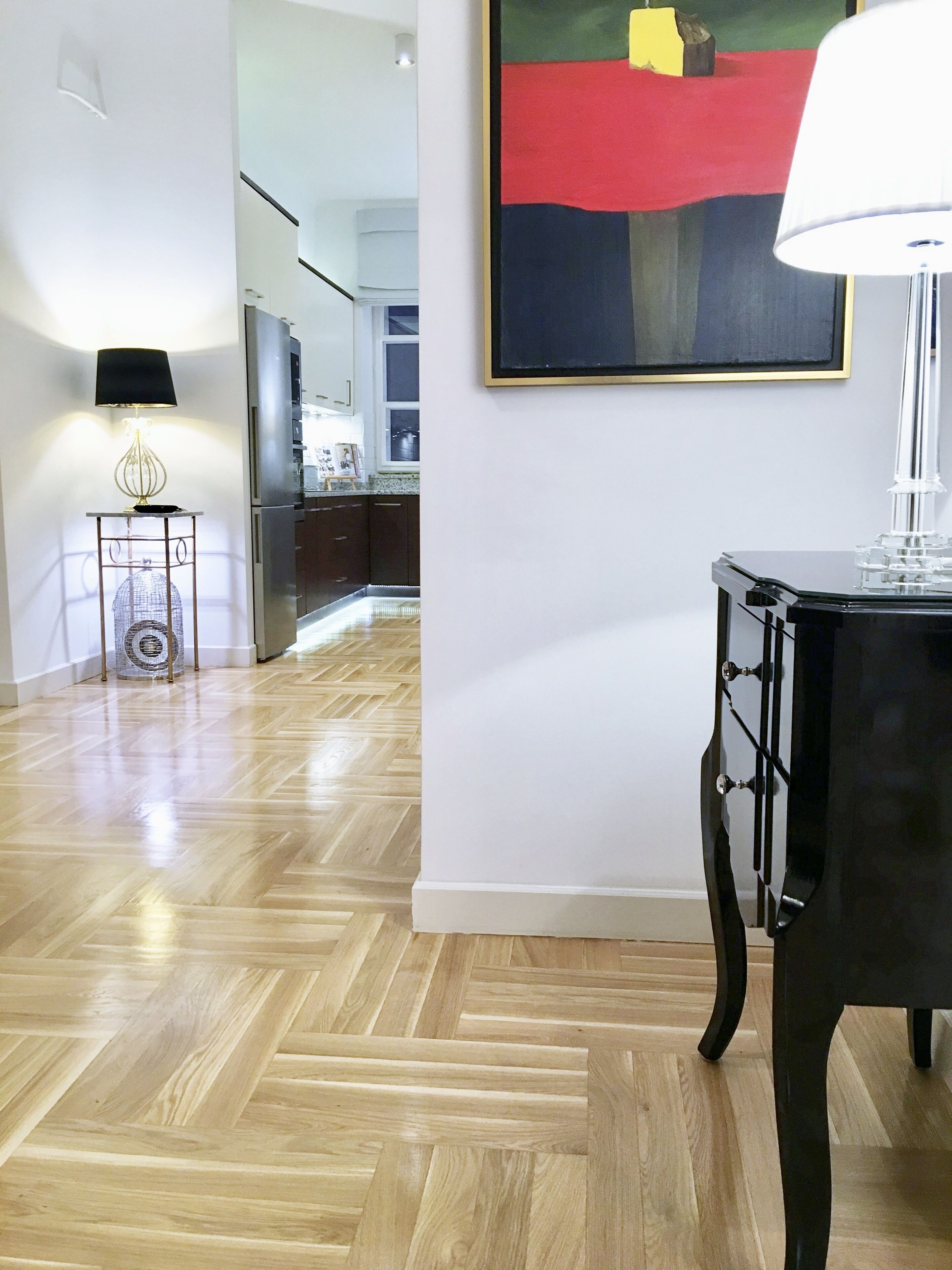 A modern interior with a herringbone wooden floor, a black table with drawers, a standing lamp, and a framed abstract painting on the wall.