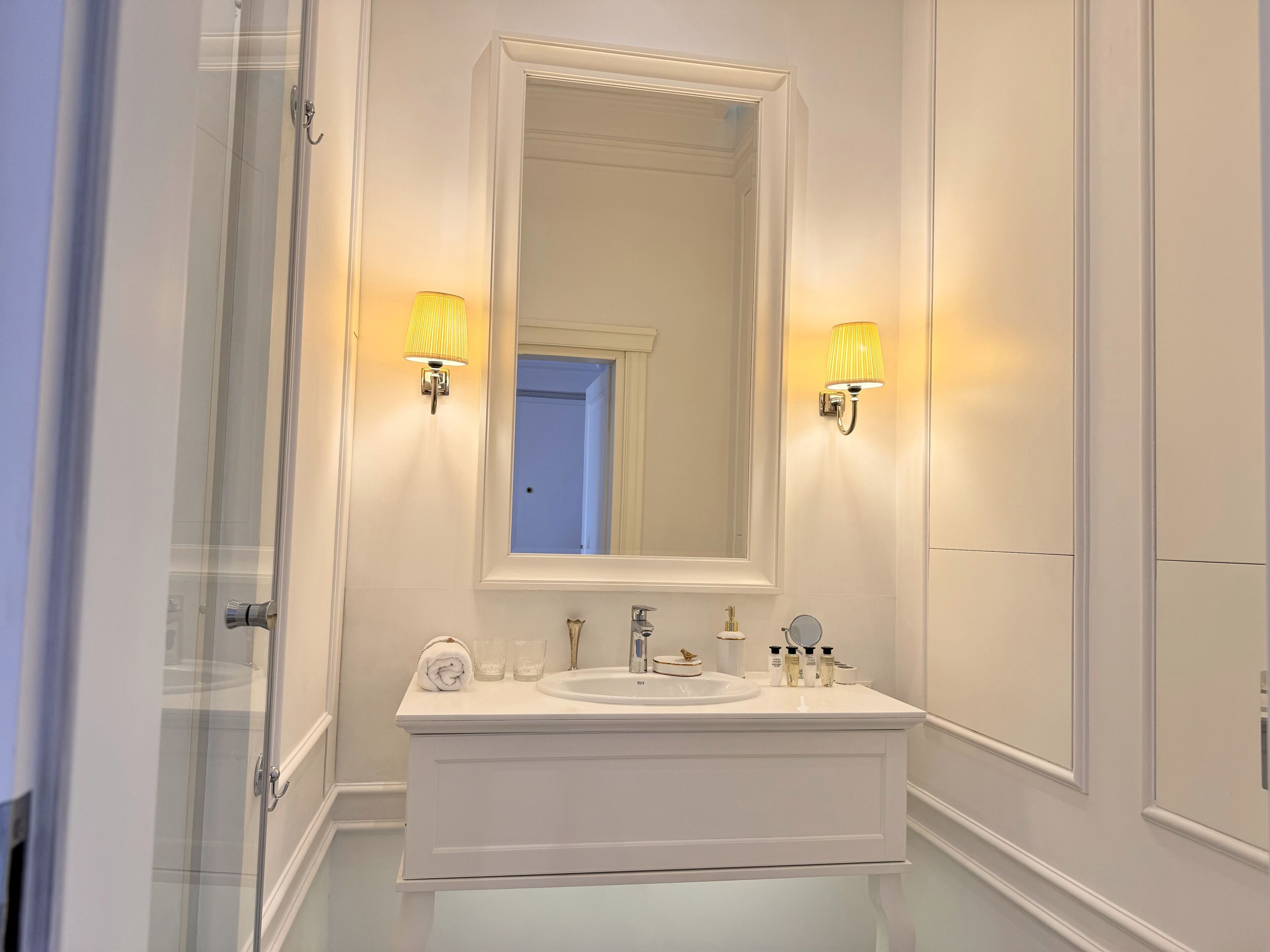 Een badkamer met een wit wastafelmeubel, een grote spiegel, twee wandlampen en een douchecabine van glas.