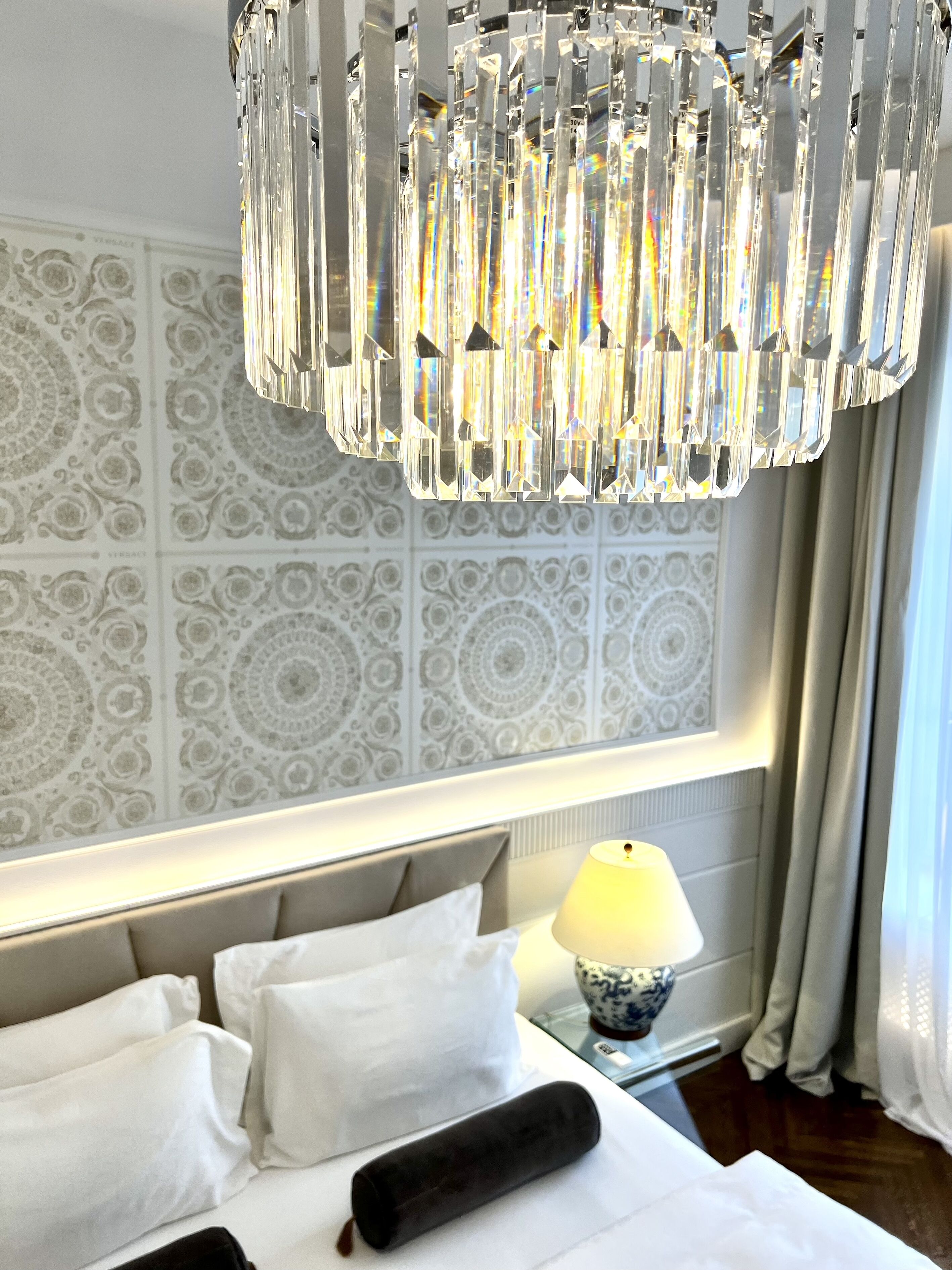 A hotel room with a large chandelier, patterned wallpaper, a bed with white linens, a bedside table with a lamp, and a window with curtains.