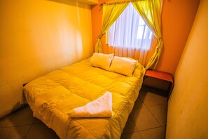 Une petite chambre avec un lit simple, une couverture jaune, un seul oreiller et un banc adossé au mur.