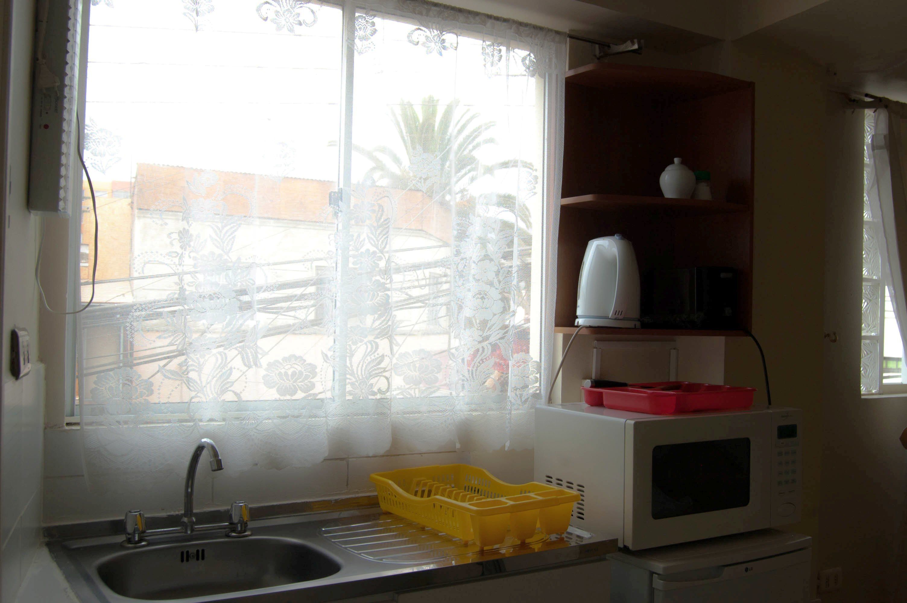Una cocina con un lavabo, un escurreplatos amarillo, un microondas blanco y un estante de madera con un jarrón blanco.