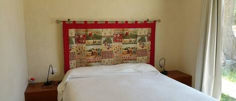 A bedroom with a bed, a red and white quilted headboard, bedside tables, and a window with curtains.