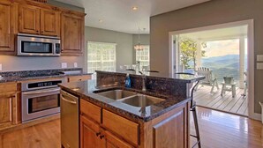 A kitchen with wooden cabinets, stainless steel appliances, a granite countertop, and a large window overlooking a deck with outdoor seating.