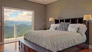 A bedroom with a large bed, two bedside lamps, a view of mountains through a large window, and a wooden floor.