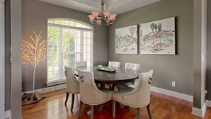 A dining room with a round table, six chairs, a chandelier, and two paintings on the wall.