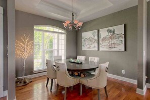 A dining room with a round table, six chairs, a chandelier, and two paintings on the wall.