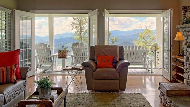 A cozy living room with a leather armchair, a red pillow, and a view of mountains through large glass doors.