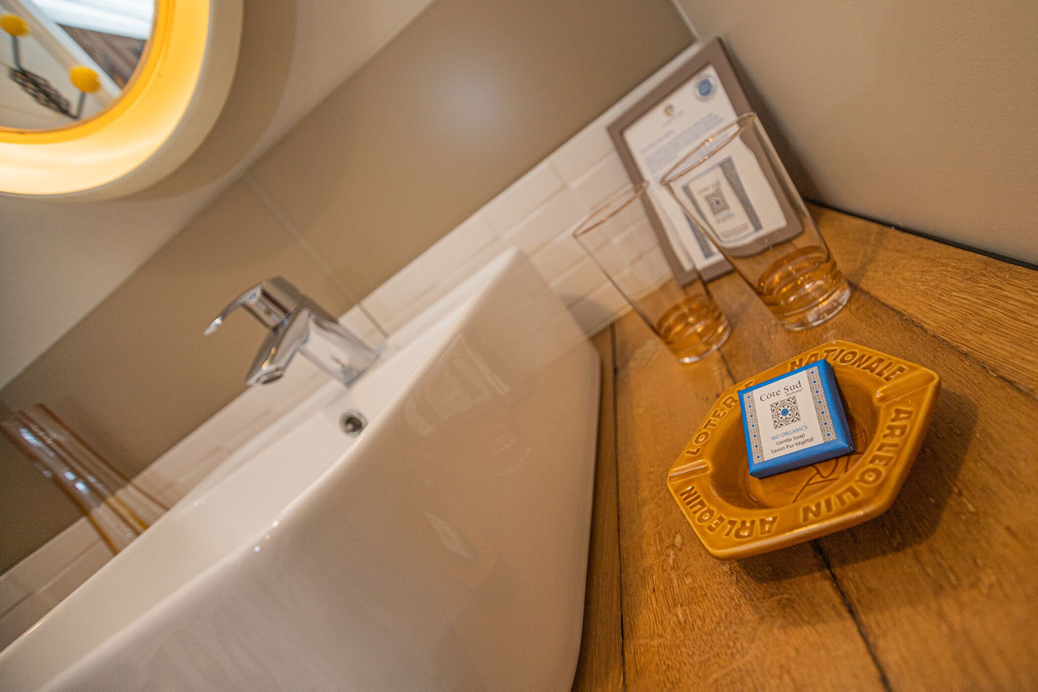 A bathroom with a bathtub, a mirror, and a wooden side table with a bottle of lotion and a glass.