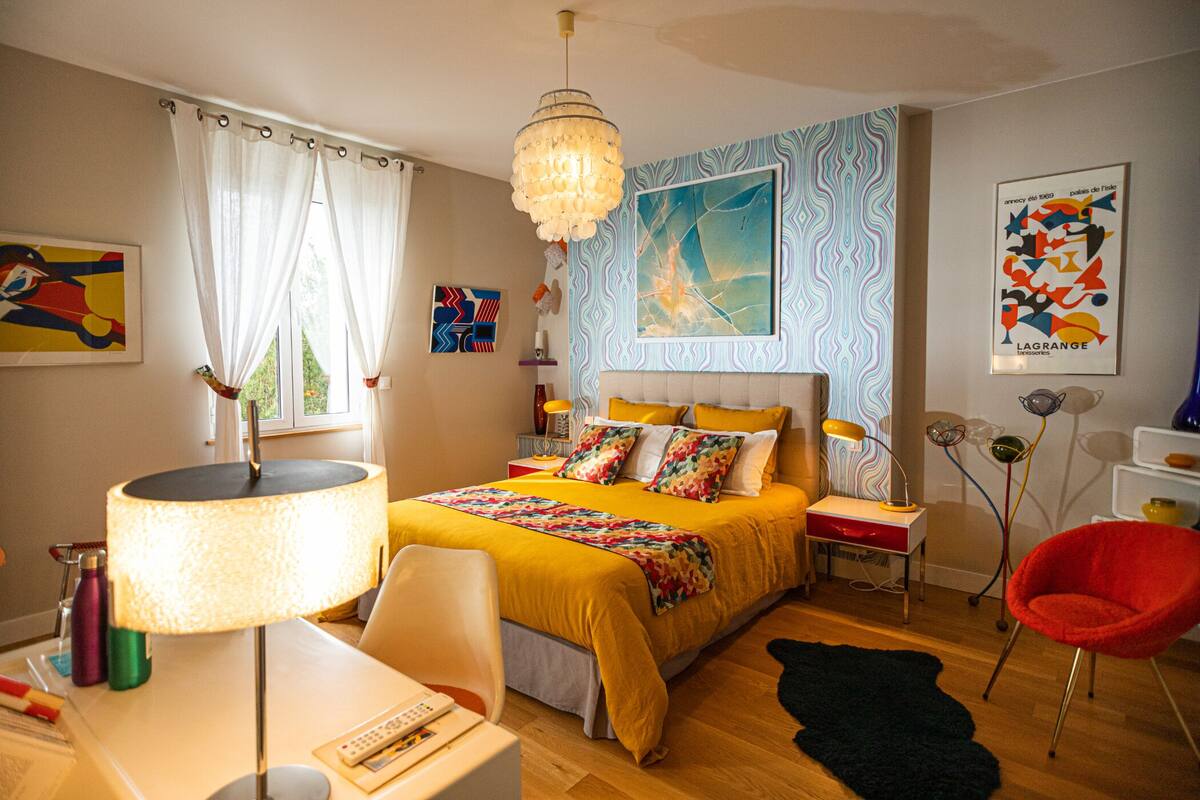 A modern bedroom with a large bed, a desk with a lamp, a red chair, and artwork on the walls.