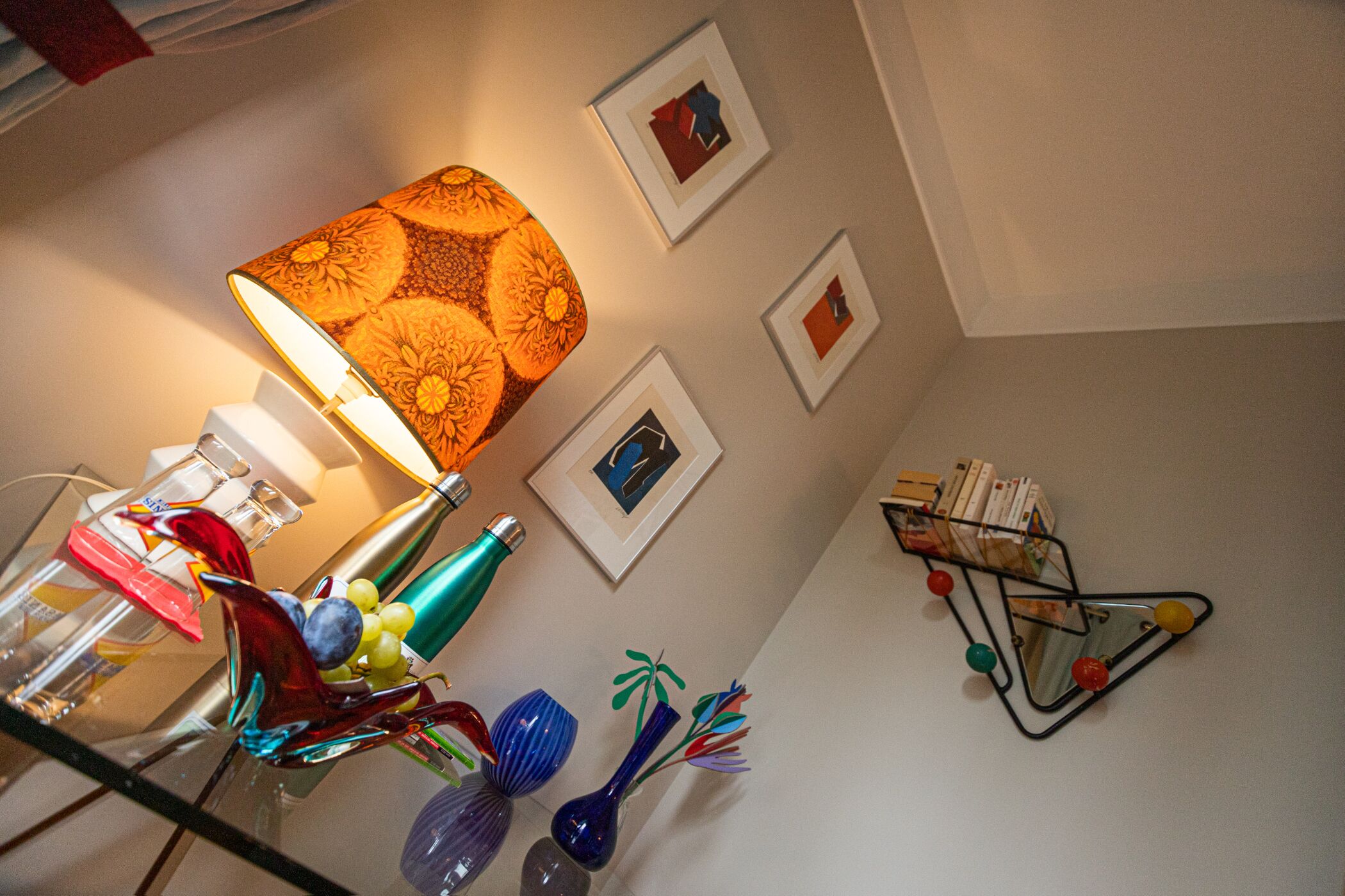 Een kamer met een lamp, afbeeldingen aan de muur en een klein boekenrek.