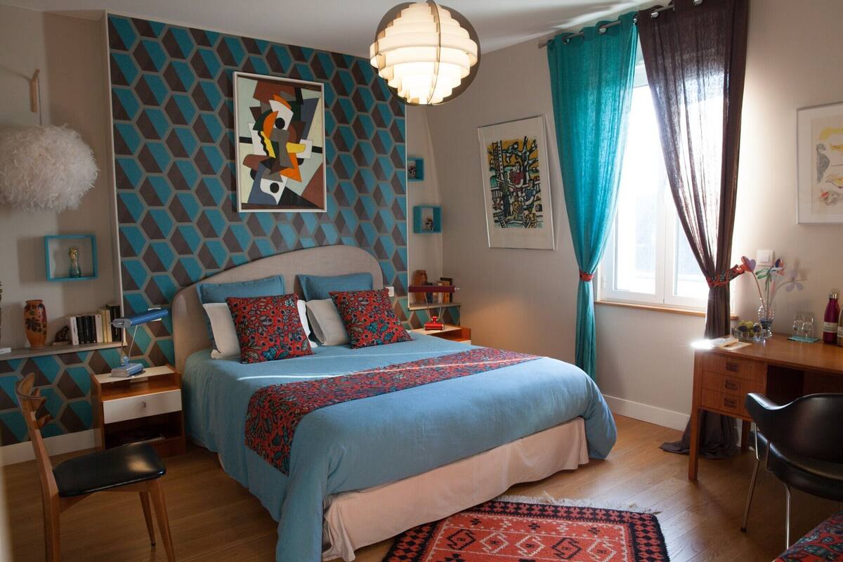 A bedroom with a bed, a desk, a chair, a lamp, and artwork on the walls.