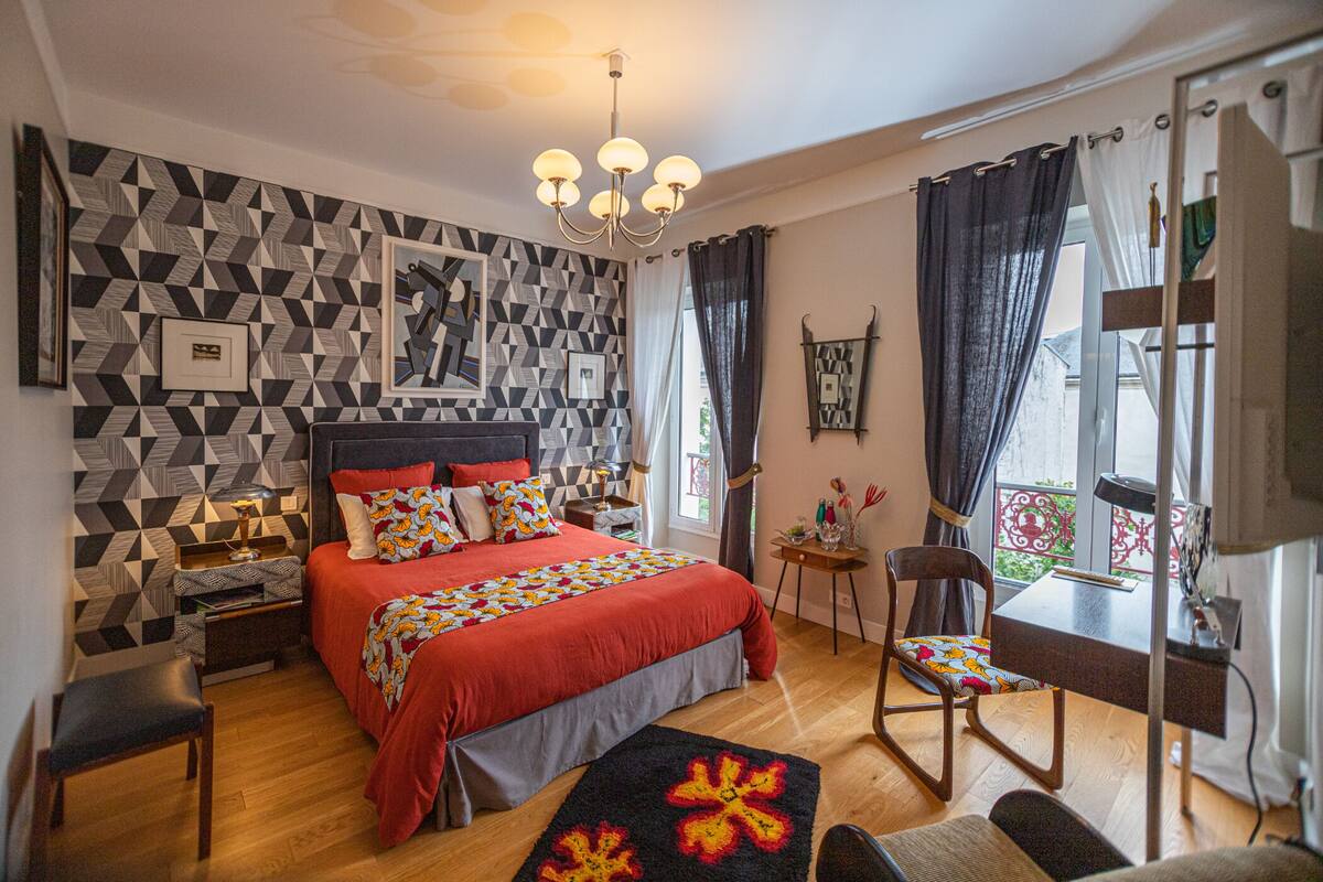 A modern bedroom with a geometric-patterned wall, a large bed with red bedding, a wooden floor, a desk with a chair, and a chandelier.