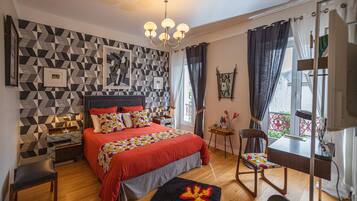 A modern bedroom with a geometric-patterned wall, a large bed with red bedding, a wooden floor, a desk with a chair, and a chandelier.