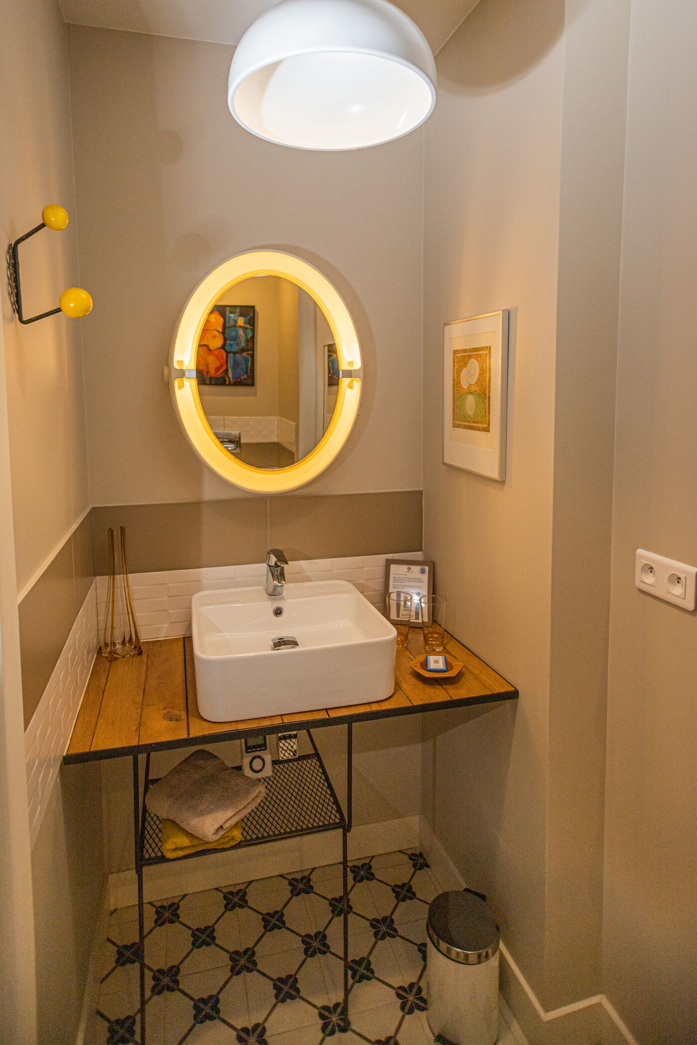A bathroom with a white sink, a mirror with a yellow light, a framed picture, and a towel on a shelf.