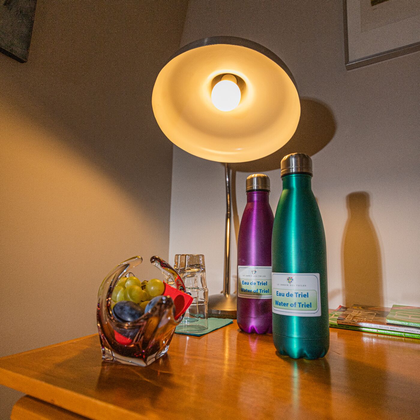 A table with a glass fruit bowl, two water bottles labeled "Eau de Triel Water of Triel," a small glass, and a lamp.