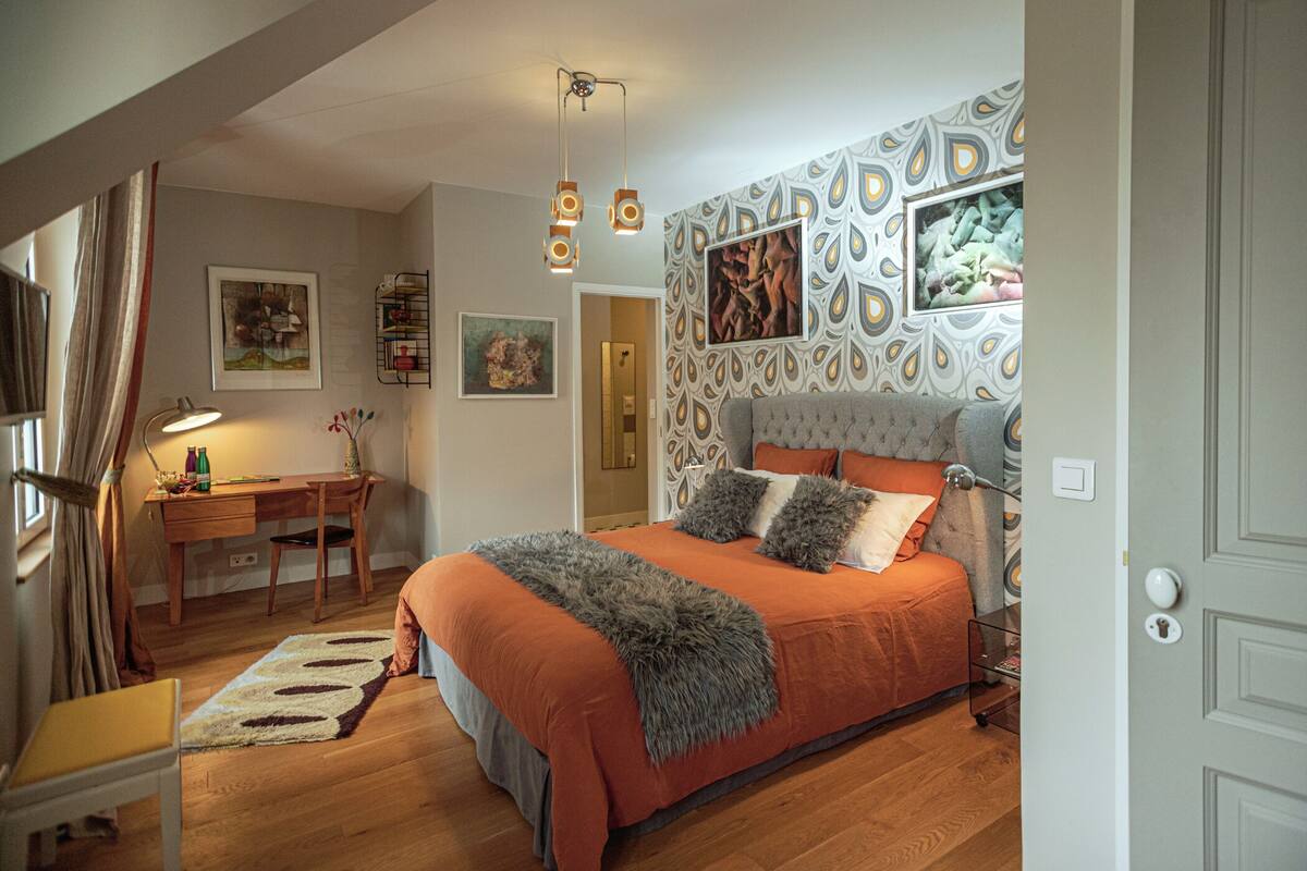 A bedroom with a bed, a desk, and a chair. There are pictures on the wall and a patterned wallpaper.