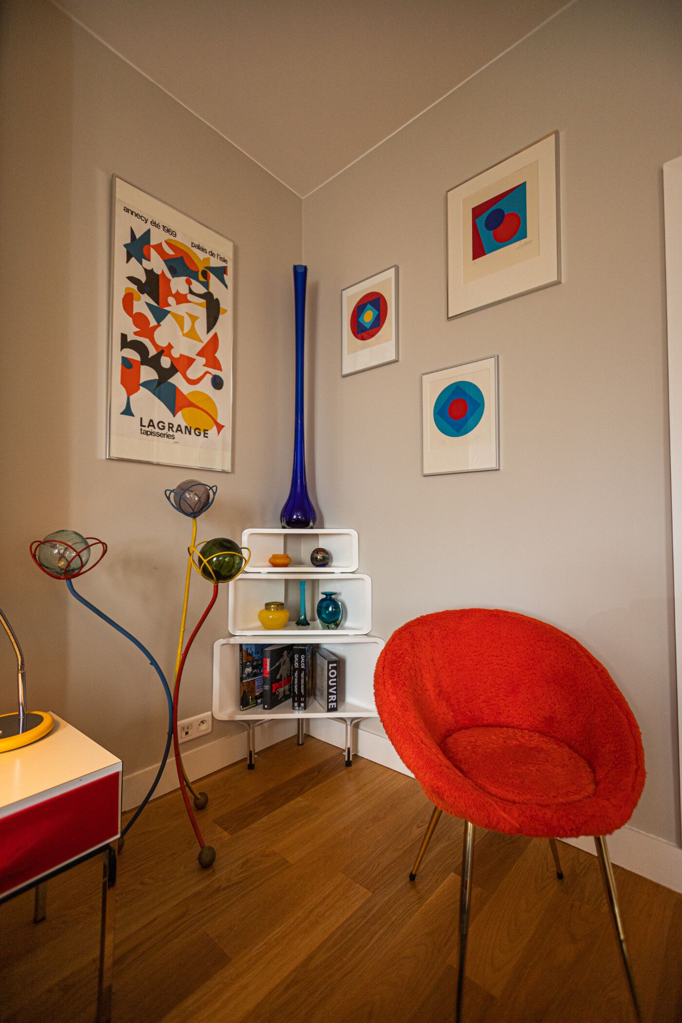 Ein Zimmer mit einem roten Sessel, einem weißen Regal mit Büchern, einer blauen Vase und abstrakter Kunst an den Wänden.