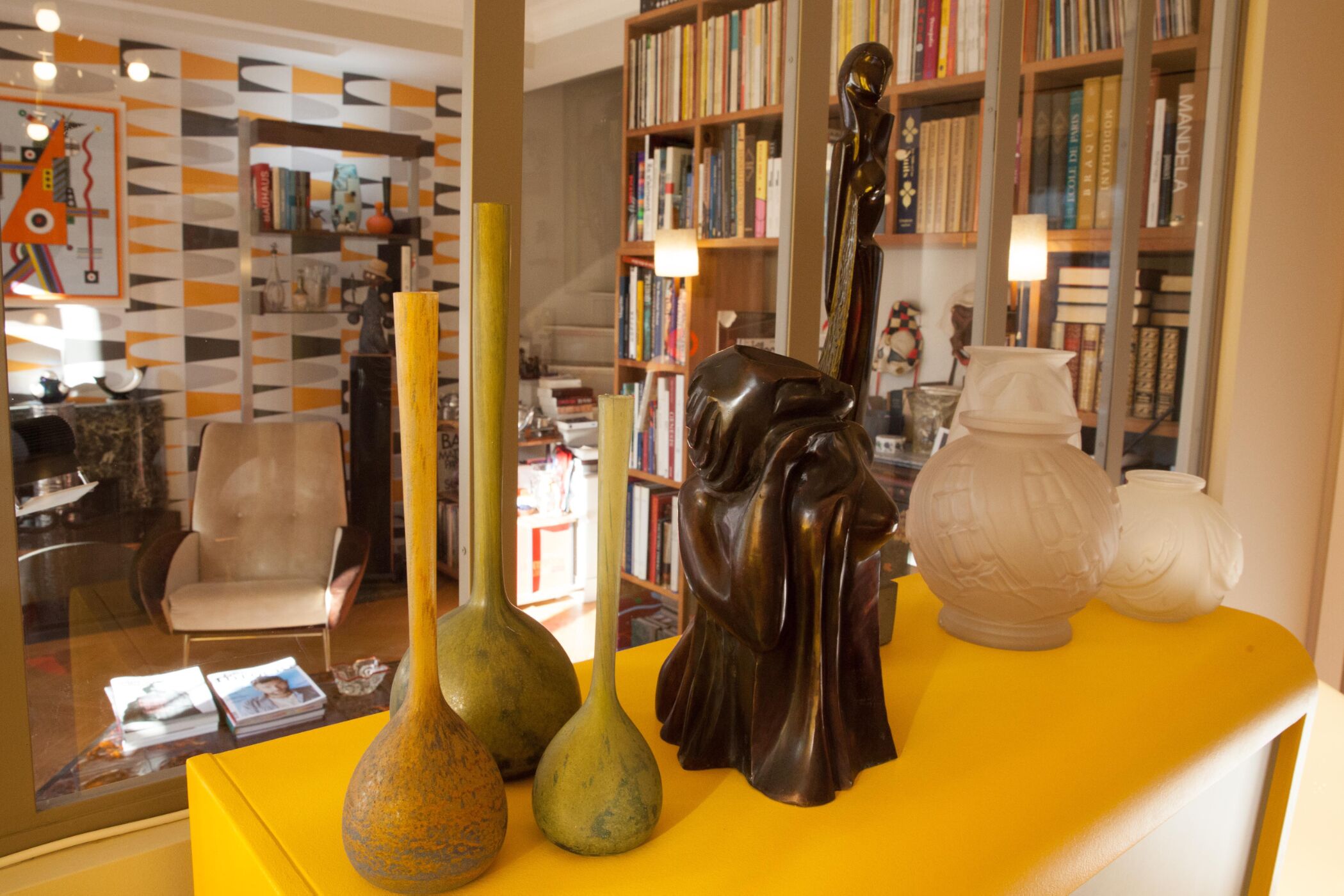 Una habitación con una mesa amarilla que exhibe varios objetos decorativos, una estantería llena de libros y una silla.
