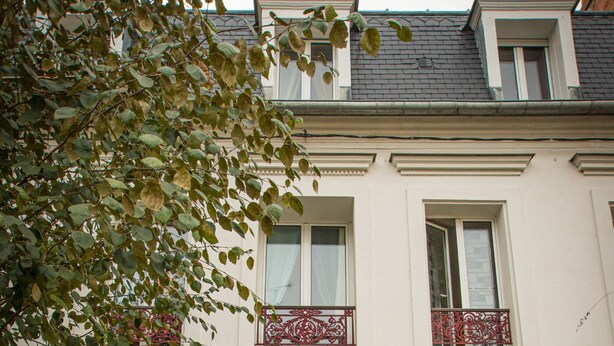 Ein zweistöckiges Gebäude mit einem zentralen Eingang, einem Balkon mit roten Geländern und Blumentöpfen rund um den Eingang.