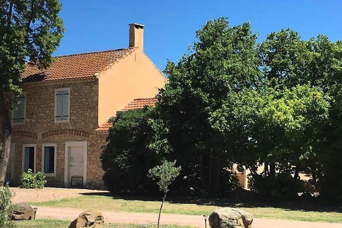 Un bâtiment en pierre, doté d’un toit en tuiles, entouré d’arbres et bordé d’une zone herbeuse parsemée de rochers.