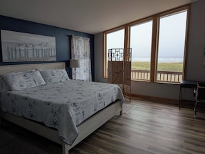 Et soveværelse med seng, et natbord, en lampe, udsigt til stranden og et stort vindue.