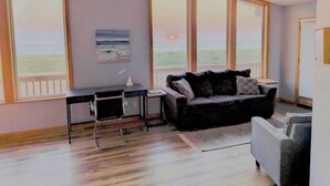 A room with a desk, a black sofa, a ceiling fan, large windows with a view of the beach, and a small rug. - Deals on Summer Sunsets! Aug 24-28, Aug 31-Sept 4. Spacious Oceanfront w/wifi (Grayland)