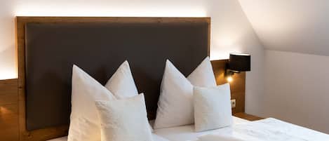 A neatly made bed with white linens and pillows, a wooden headboard, and a wall-mounted light fixture.