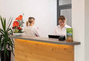 A reception desk with a wooden counter, a potted plant with red flowers, and a brochure on the counter. - Hotel Gasthof Rosenwirt (Au in der Hallertau)