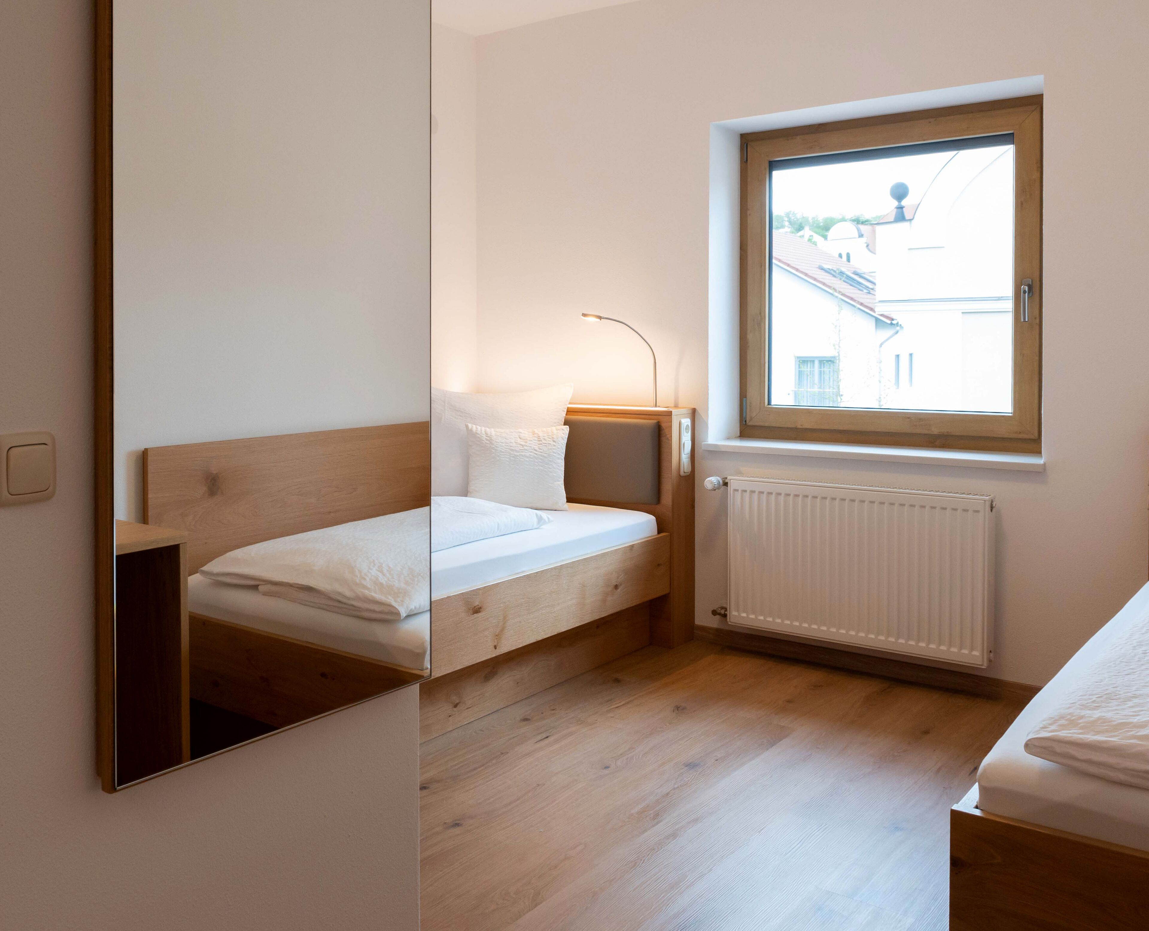 Een kleine, goed verlichte slaapkamer met een houten bed, een nachtkastje, een raam met uitzicht op gebouwen, en een radiator.