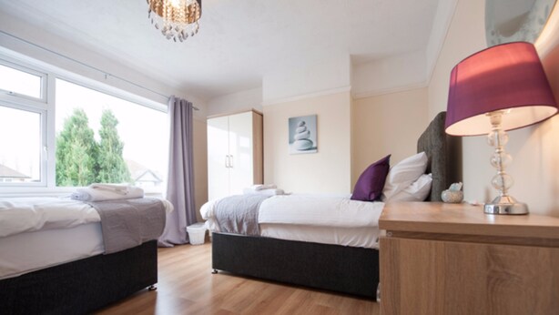 Quarto moderno com duas camas, piso de madeira, janela com cortinas e abajur.