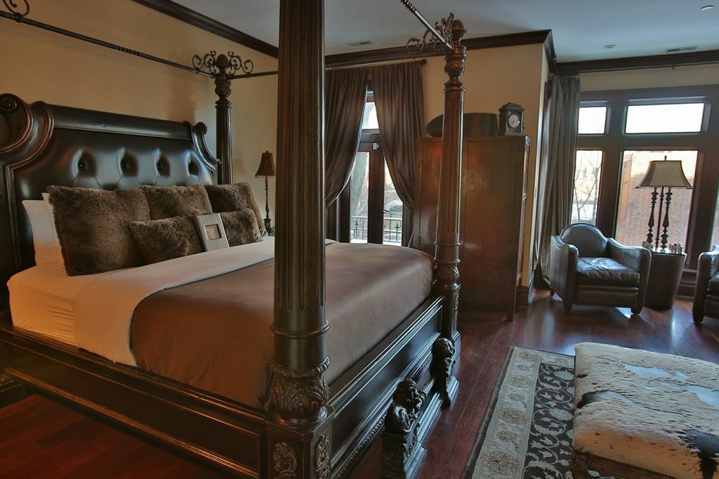 A four-poster bed with a dark wooden frame and a tufted headboard, a large window with curtains, a leather armchair, and a wooden nightstand with a lamp.