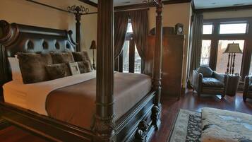 A four-poster bed with a dark wooden frame and a tufted headboard, a large window with curtains, a leather armchair, and a wooden nightstand with a lamp.