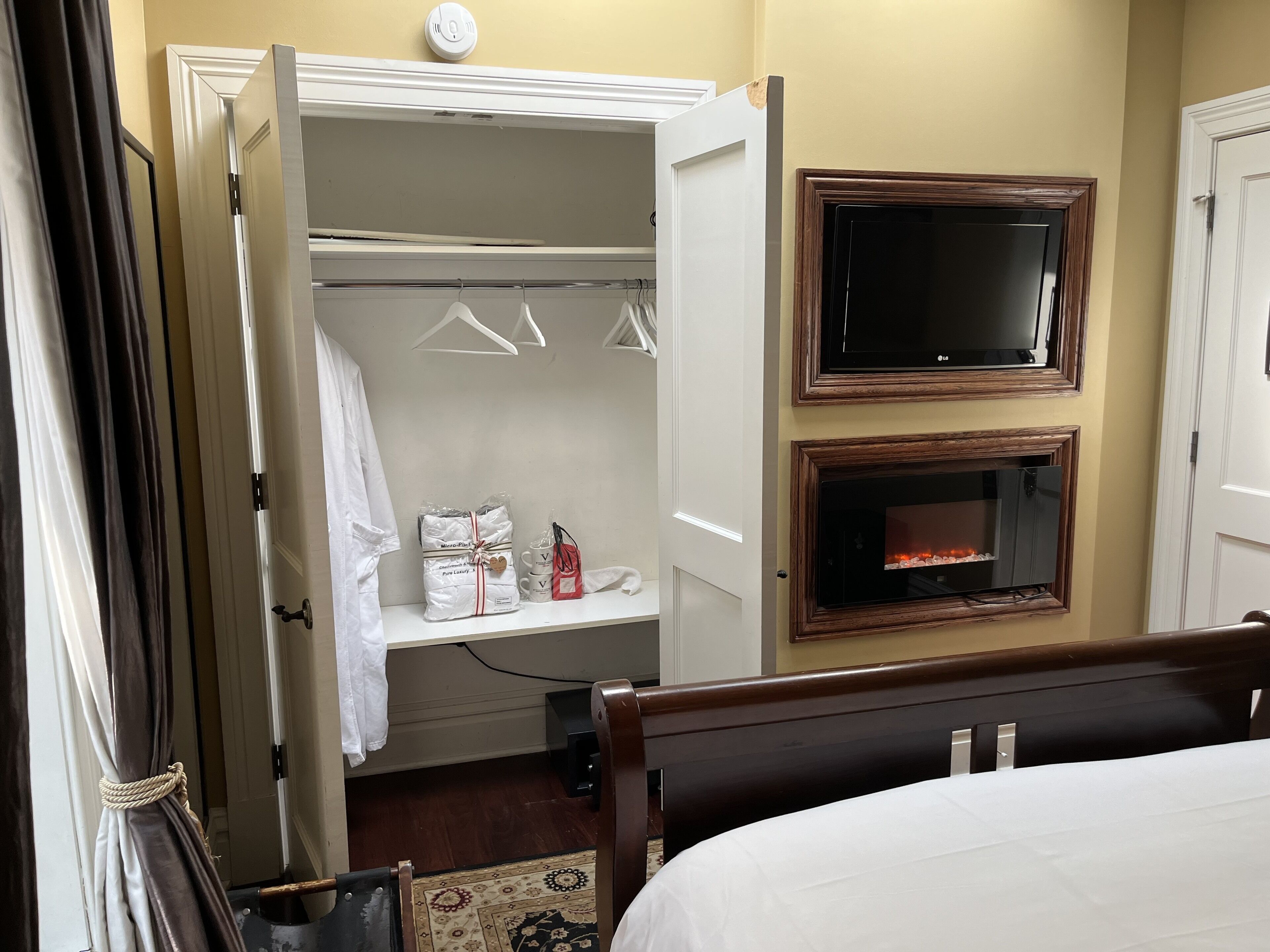 A hotel room with a bed, a TV mounted above a fireplace, a closet with hanging clothes, and a shower area.