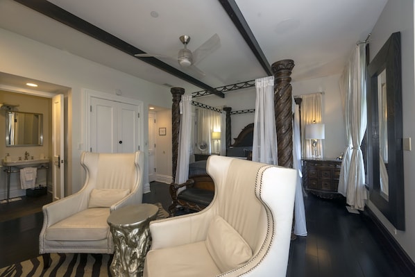 A spacious room with a large bed, a sitting area with a chair and a small table, a bathroom with a sink and mirror, and a staircase with a view of the bedroom.