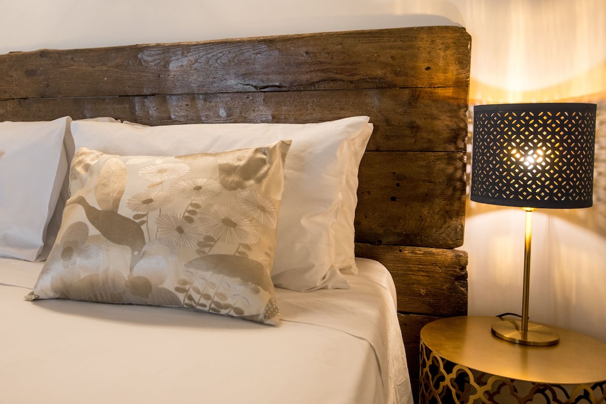 A bed with white linens and patterned pillows, a wooden headboard, and a round side table with a lamp.