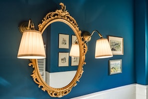 Um banheiro com um espelho grande e ricamente decorado, luminárias fixadas na parede e quadros em uma parede azul.
