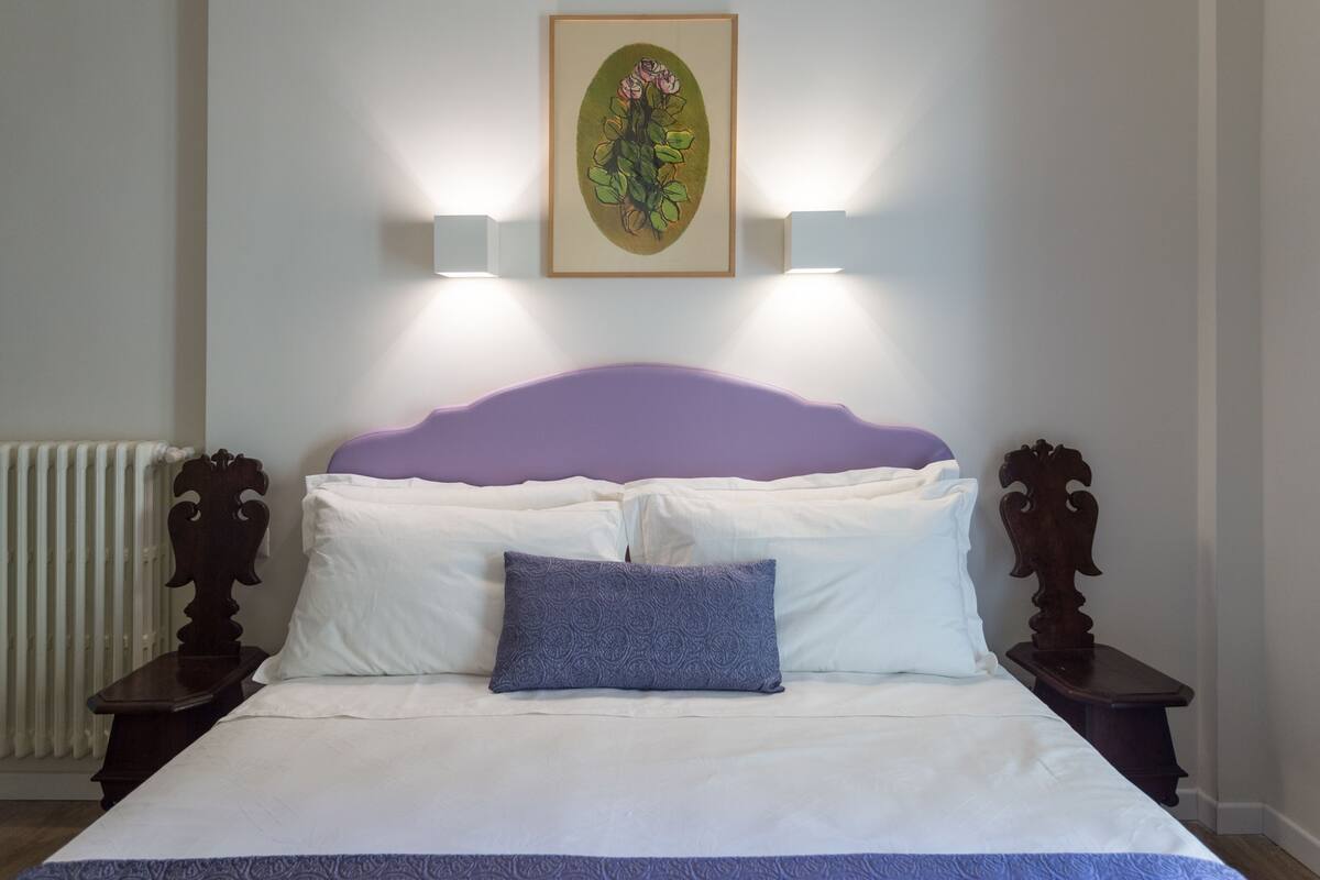 A neatly made bed with white and purple bedding, two bedside tables with lamps, and a framed artwork above the bed.