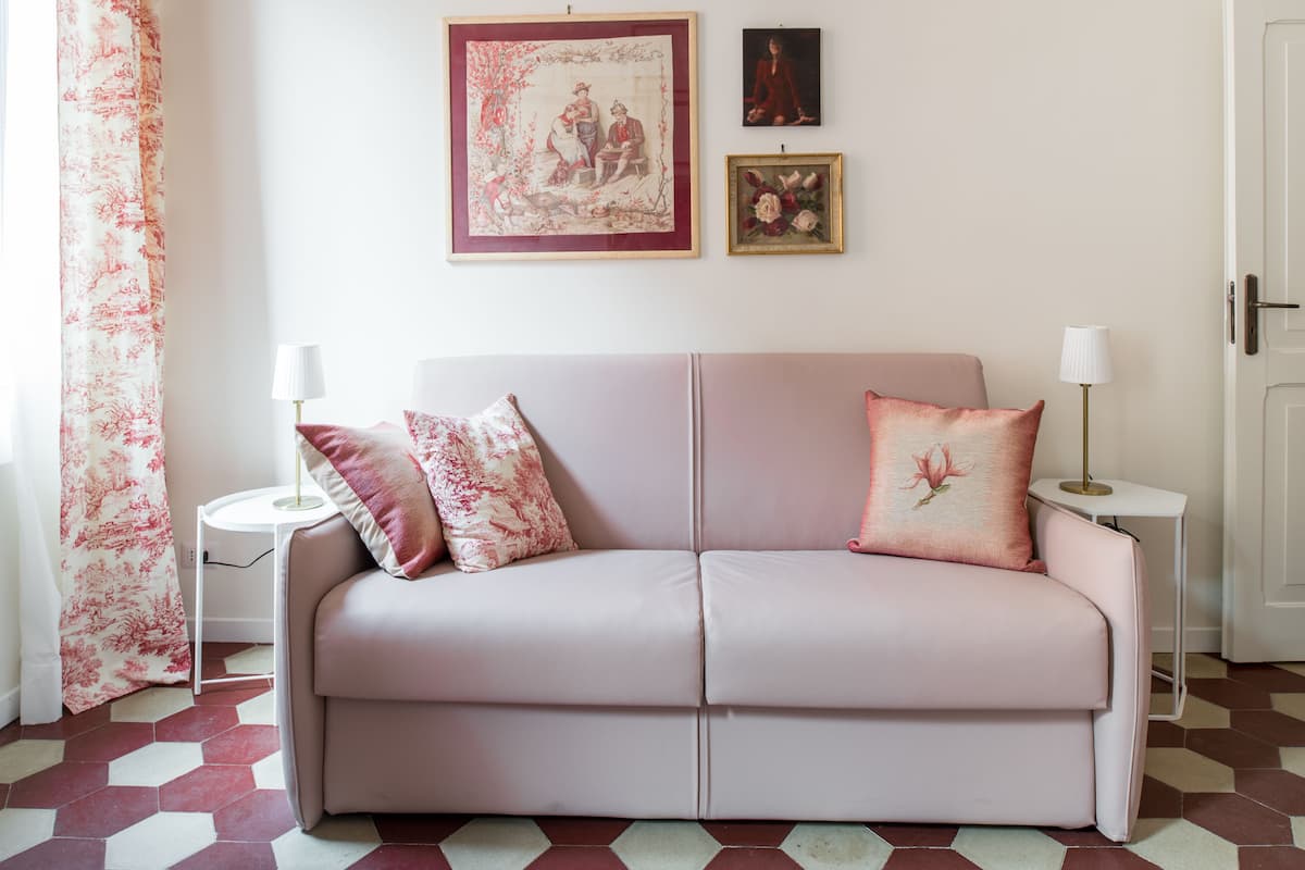 A room with a pink sofa, two pillows, a framed picture, a small table with a lamp, and a door.
