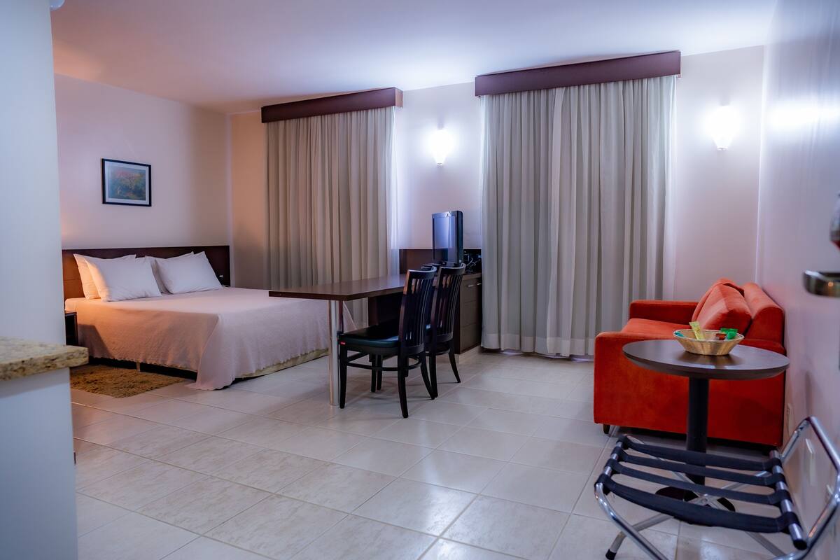 A hotel room with a bed, a desk with a chair, a small table with a bowl, and a red sofa.