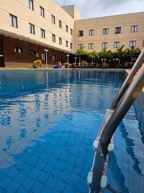 A swimming pool with tiled flooring, surrounded by a building with multiple windows and a flat roof. - Hotel Santos Dumont (Goiania)