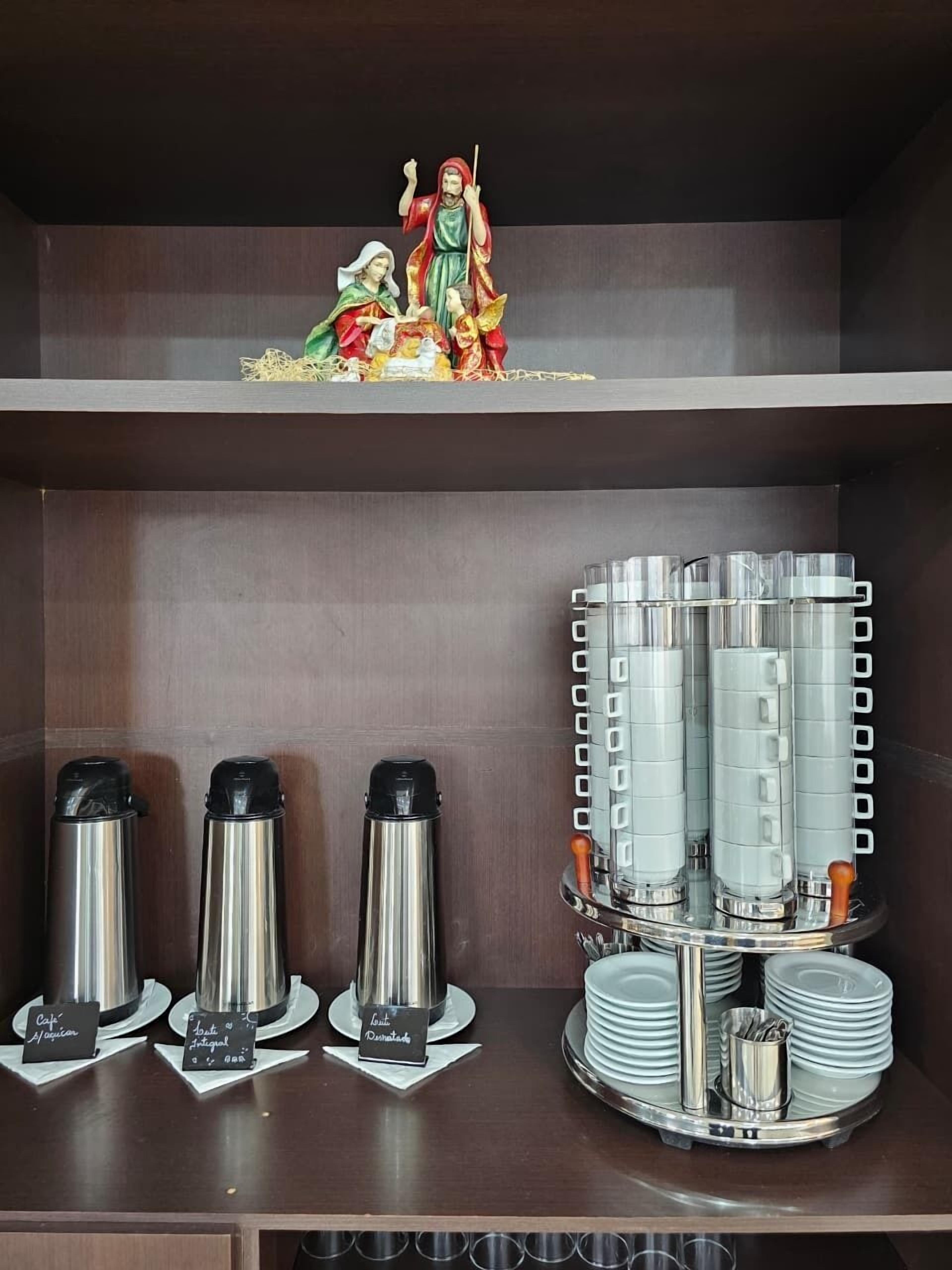 A hotel room with a shelf displaying a religious statue, a coffee station with multiple coffee makers, and a tray with stacked plates and a cup.
