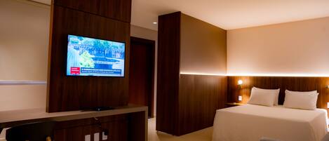 Una habitación de hotel con un escritorio de madera, un televisor en la pared, una cama con ropa de cama blanca y una mesita de noche con lámpara.