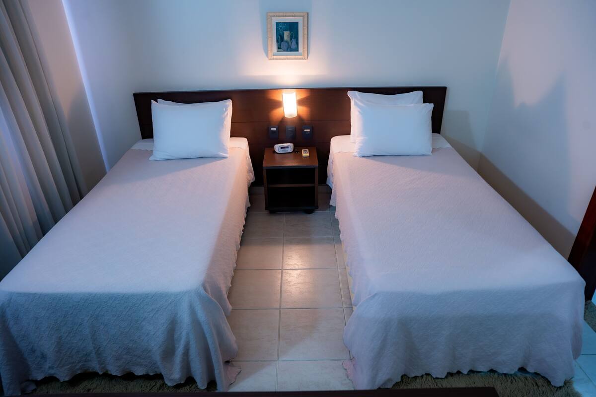 A hotel room with two single beds, a nightstand with a lamp, and a framed picture on the wall.