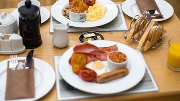 A breakfast table with plates of eggs, bacon, tomatoes, beans, and toast. There are also condiments, a coffee pot, and a glass of orange juice.