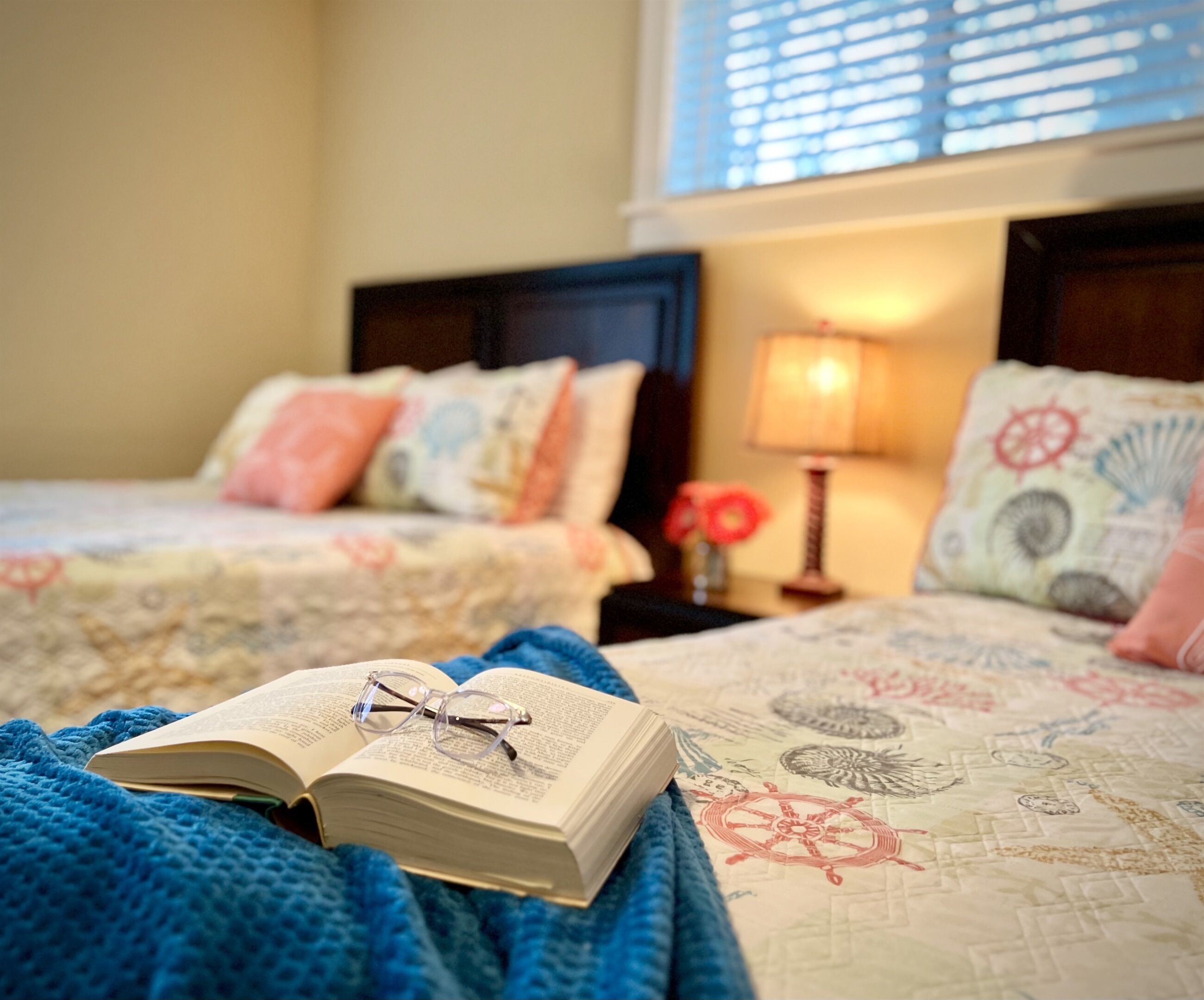 Quarto de hotel com duas camas, um cobertor azul, um livro aberto e uma luminária.