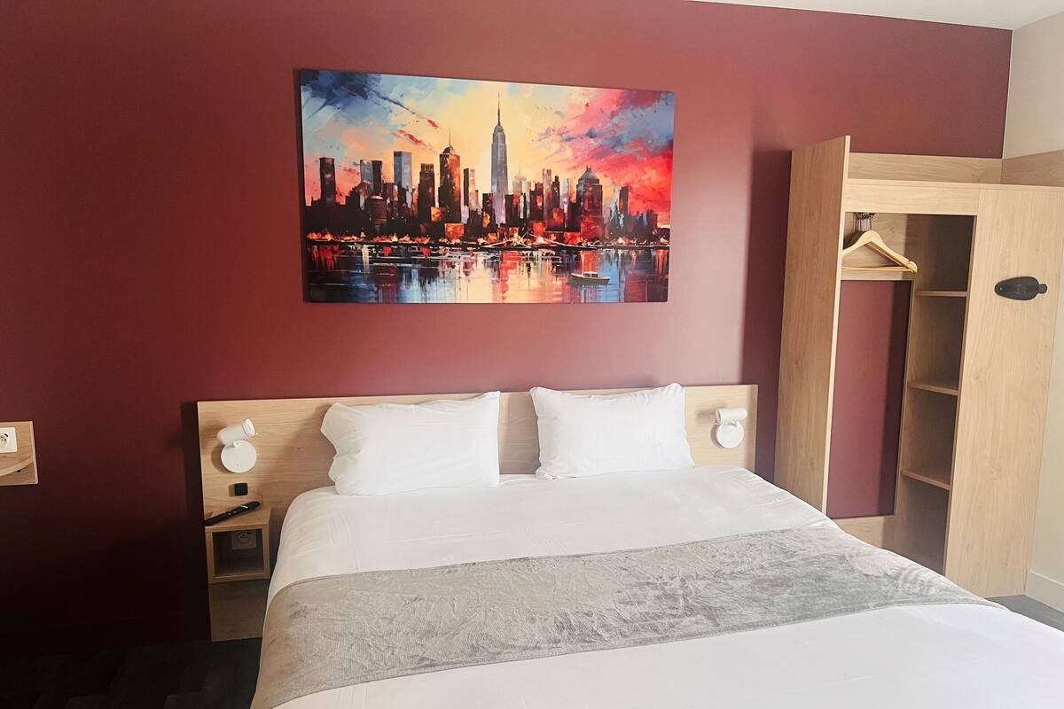 Een hotelkamer met een bed, een nachtkastje, een houten garderobe en een stadsgezicht schilderij aan de muur.