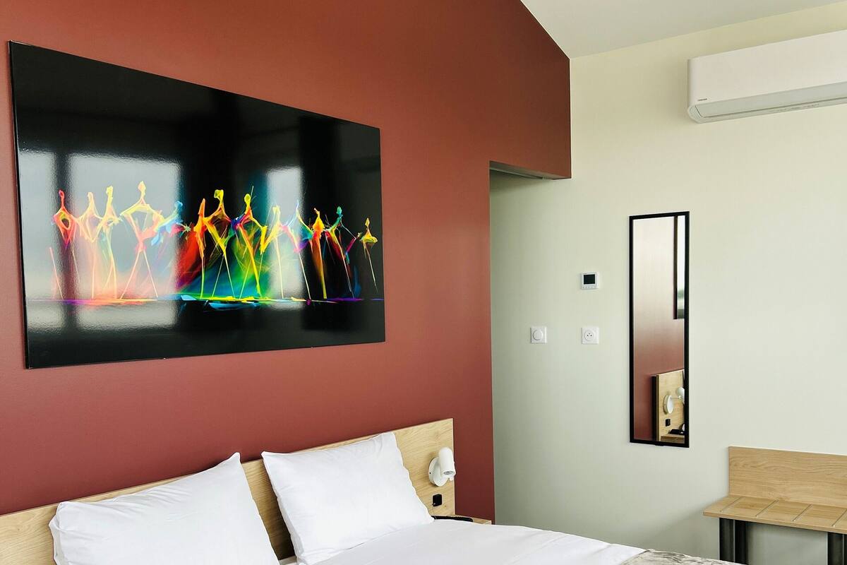 Een hotelkamer met een groot bed, een nachtkastje, een wandlamp, een spiegel en een kleurrijk kunstwerk aan de muur.