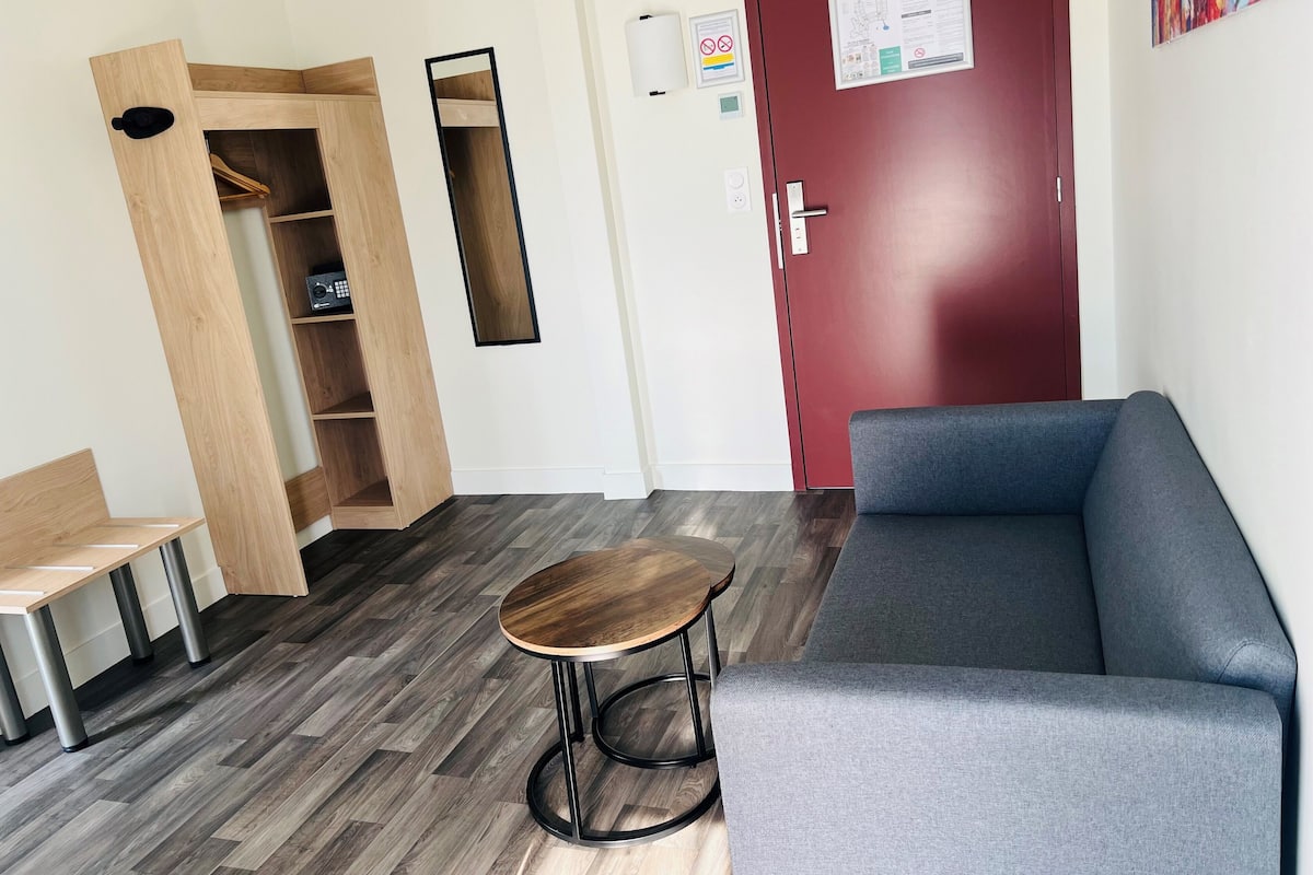 Een moderne kamer met een grijze bank, een houten bankje, een klein rond tafeltje en een garderobe met planken.